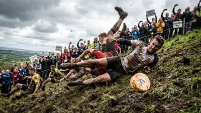 Several muddy men tumble down a steep, grassy hill in pursuit of a rolling cheese wheel, as spectators cheer from the side.