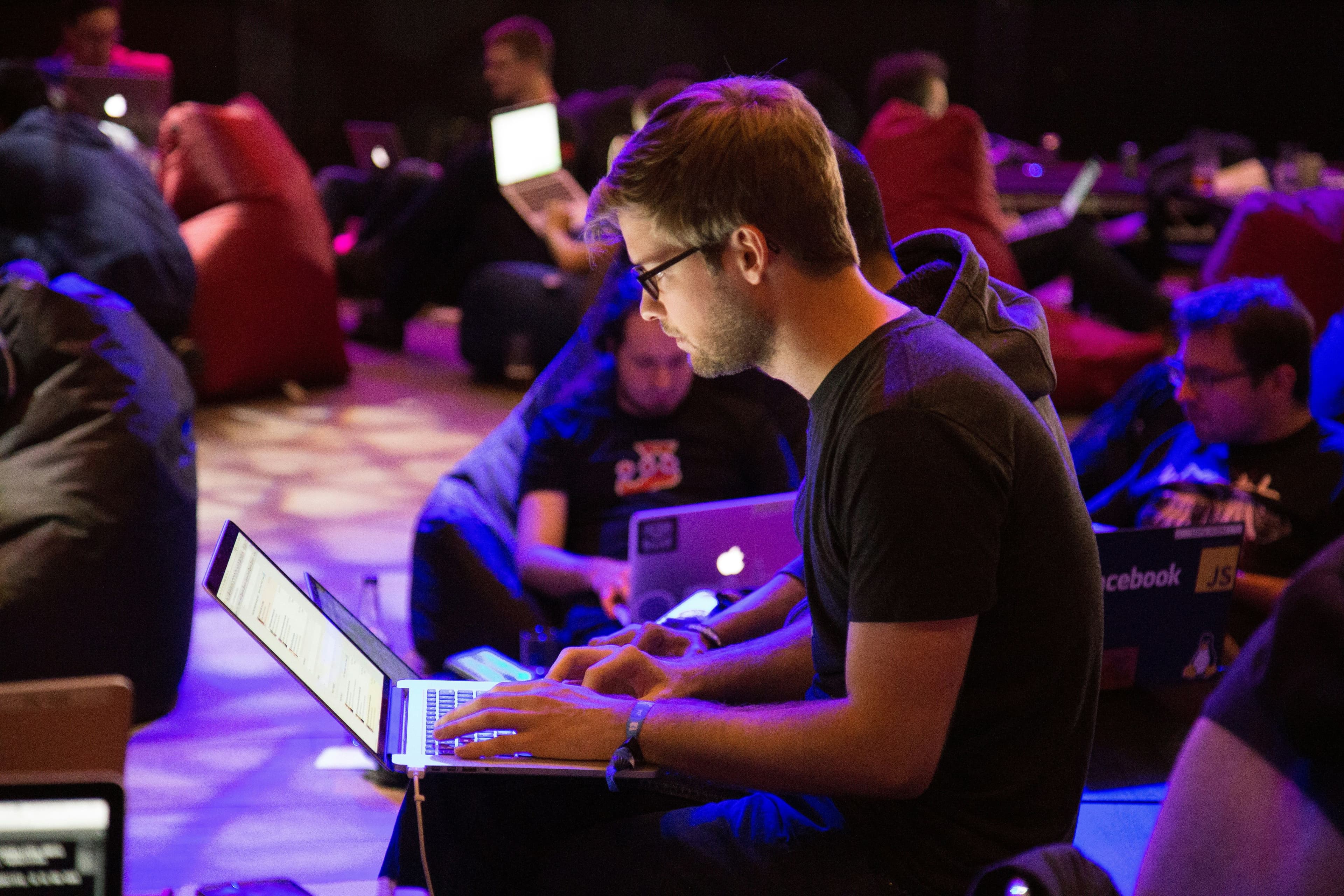 Man in glasses typing on a laptop among other attendees working on laptops in a dimly lit hall
