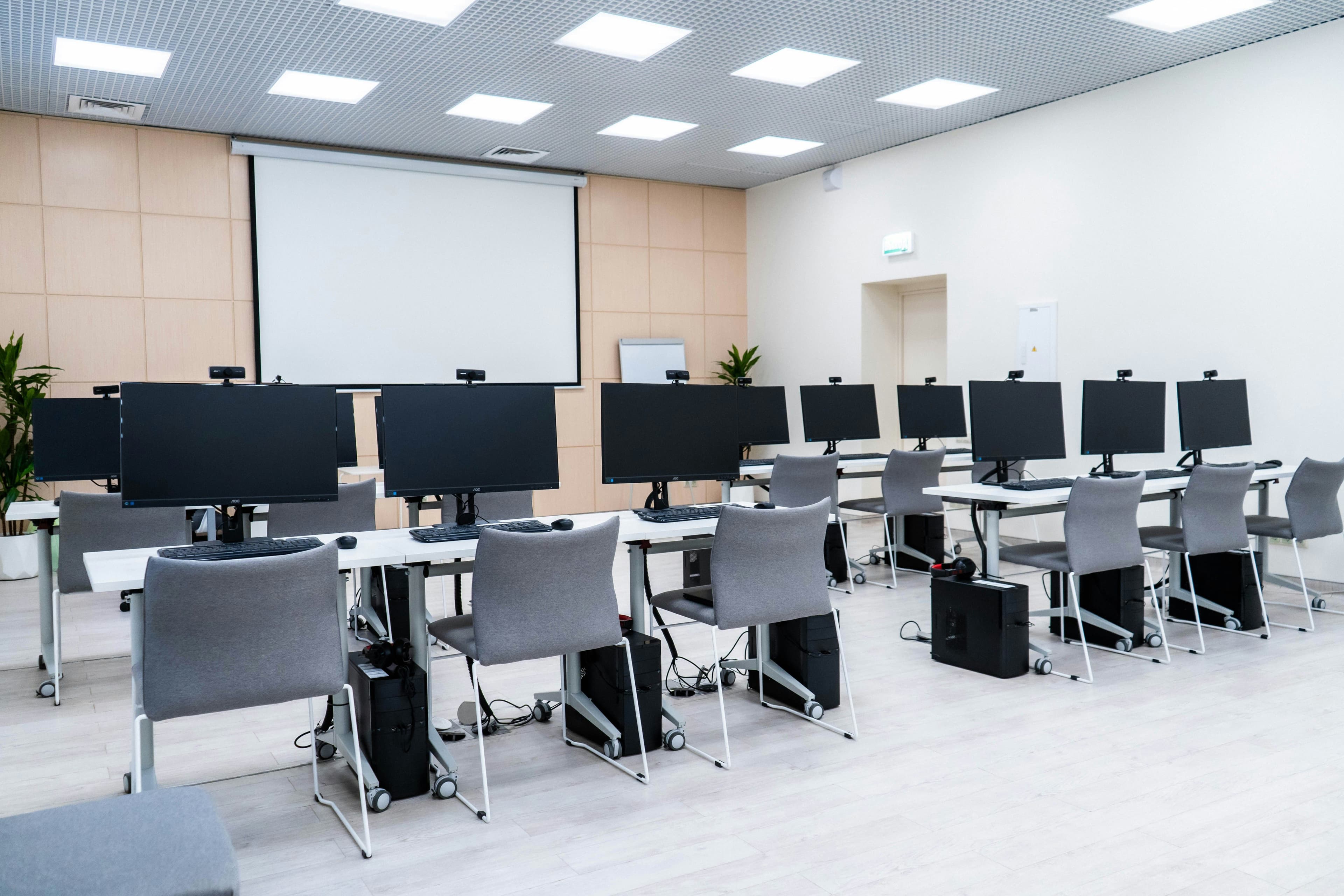 Rows of desks with black monitors, webcams and grey chairs in an empty training room facing a projector screen