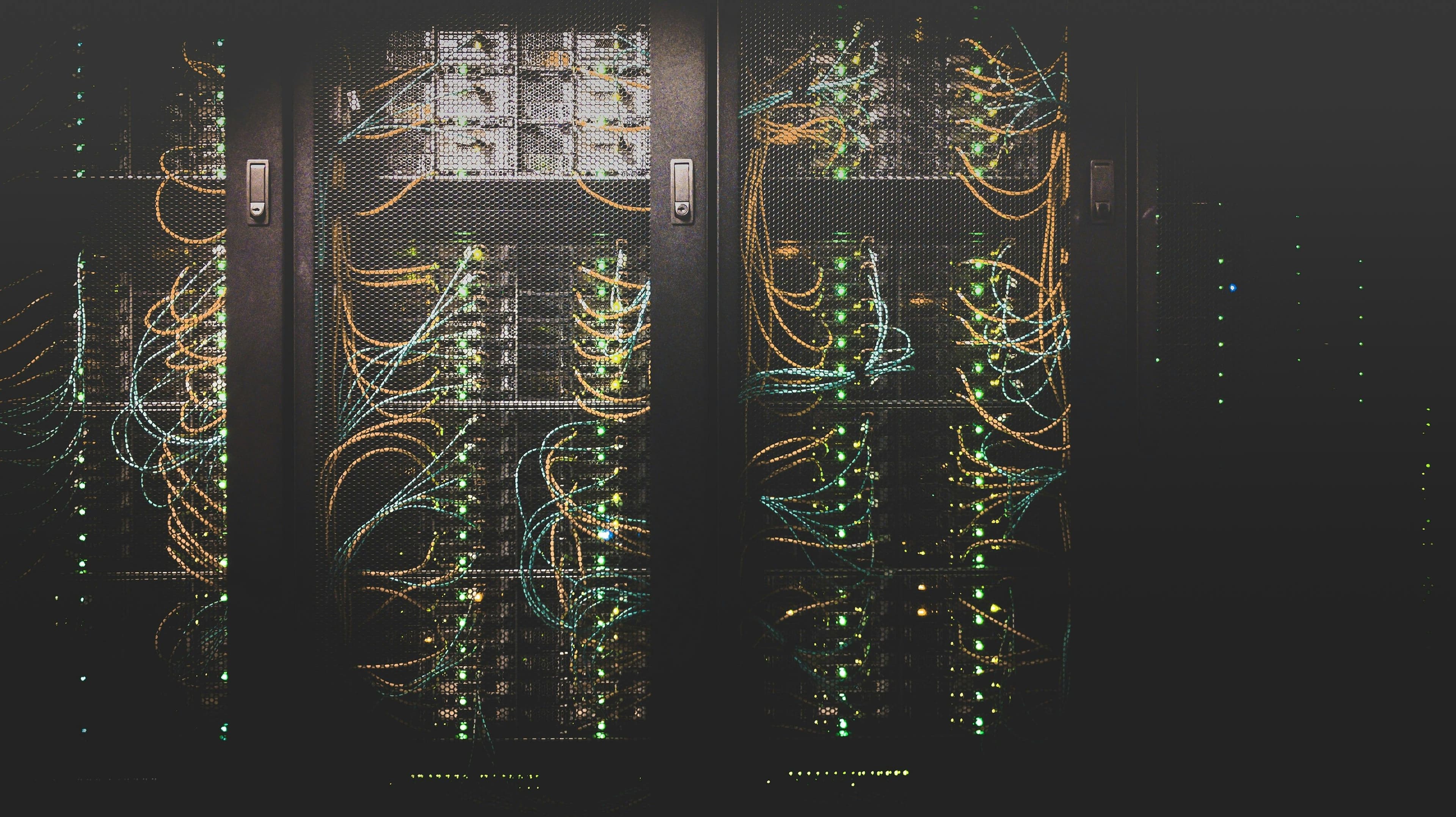Rows of dark server racks with glowing green lights and orange and blue cables fanned across the mesh doors of a data centre