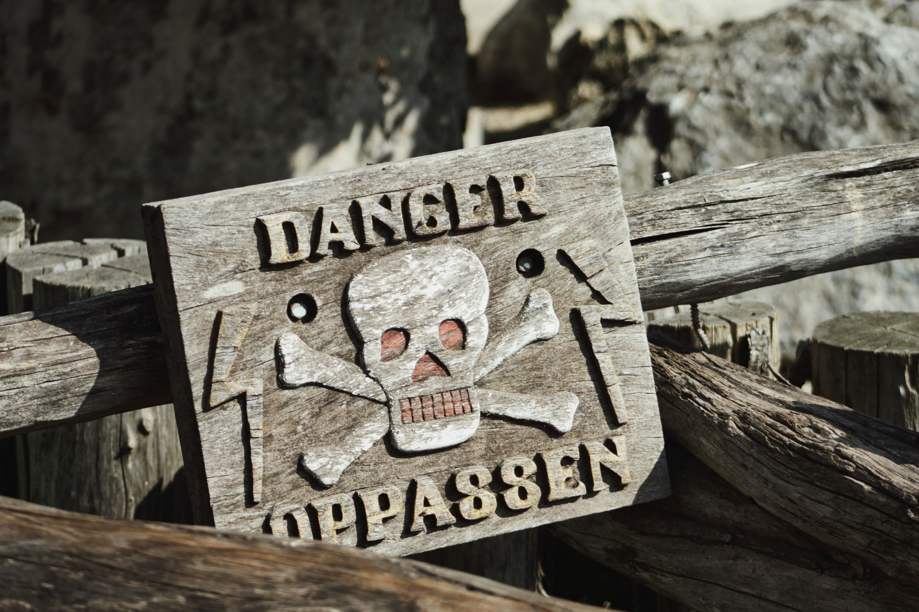 Weathered wooden sign with carved skull and crossbones above the words "DANGER" and "OPPASSEN" on a rustic fence