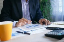 Person in a dark blazer reviewing a bar chart on a tablet beside a calculator, notepad and pen on a desk