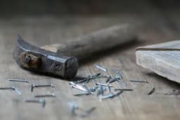 An old iron hammer and scattered steel nails lying on a weathered wooden workbench, with a piece of timber at right