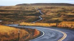 A winding two-lane road curving through dry golden grassland and rolling hills under an overcast sky