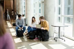 Three students sit together on a bench in a sunlit atrium, one reviewing notes on a laptop while others look on