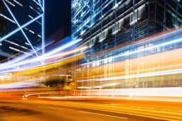 Long-exposure night photo of light trails streaking past glass office towers on a city street