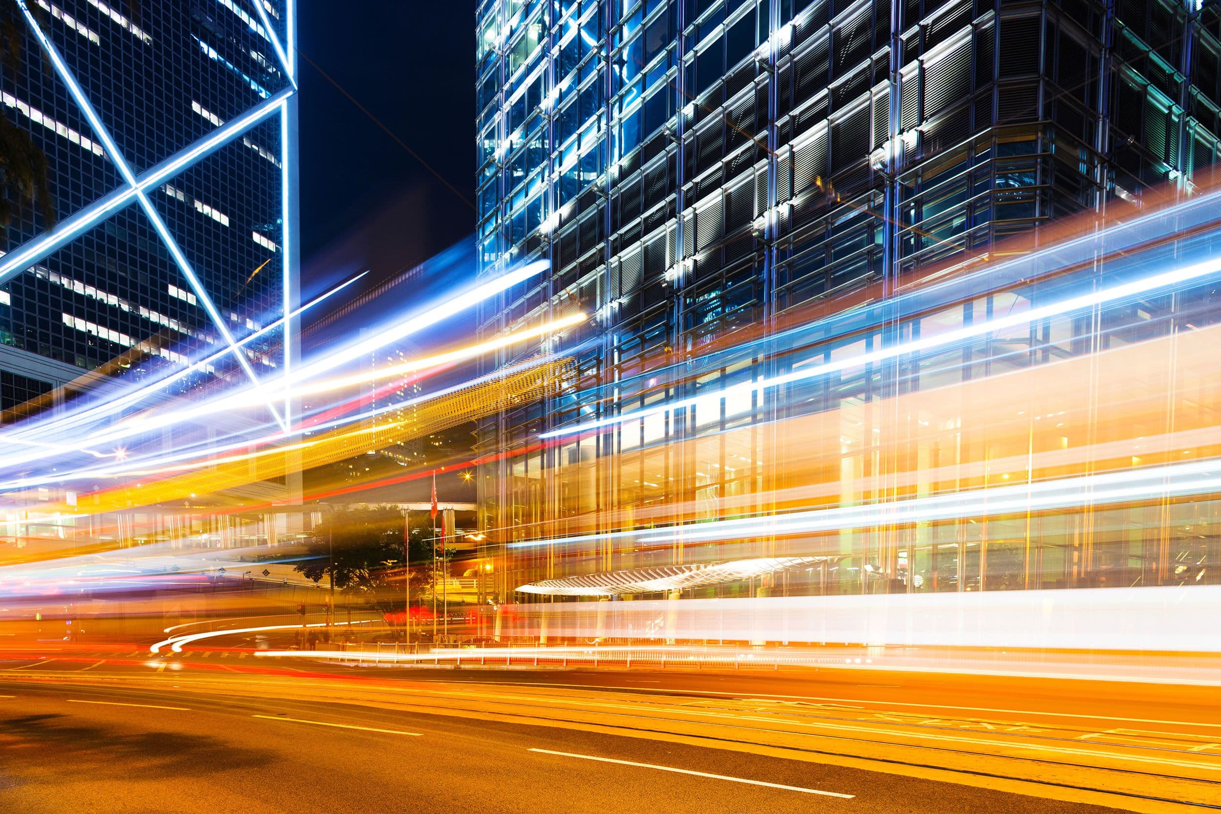 Long-exposure night photo of light trails streaking past glass office towers on a city street