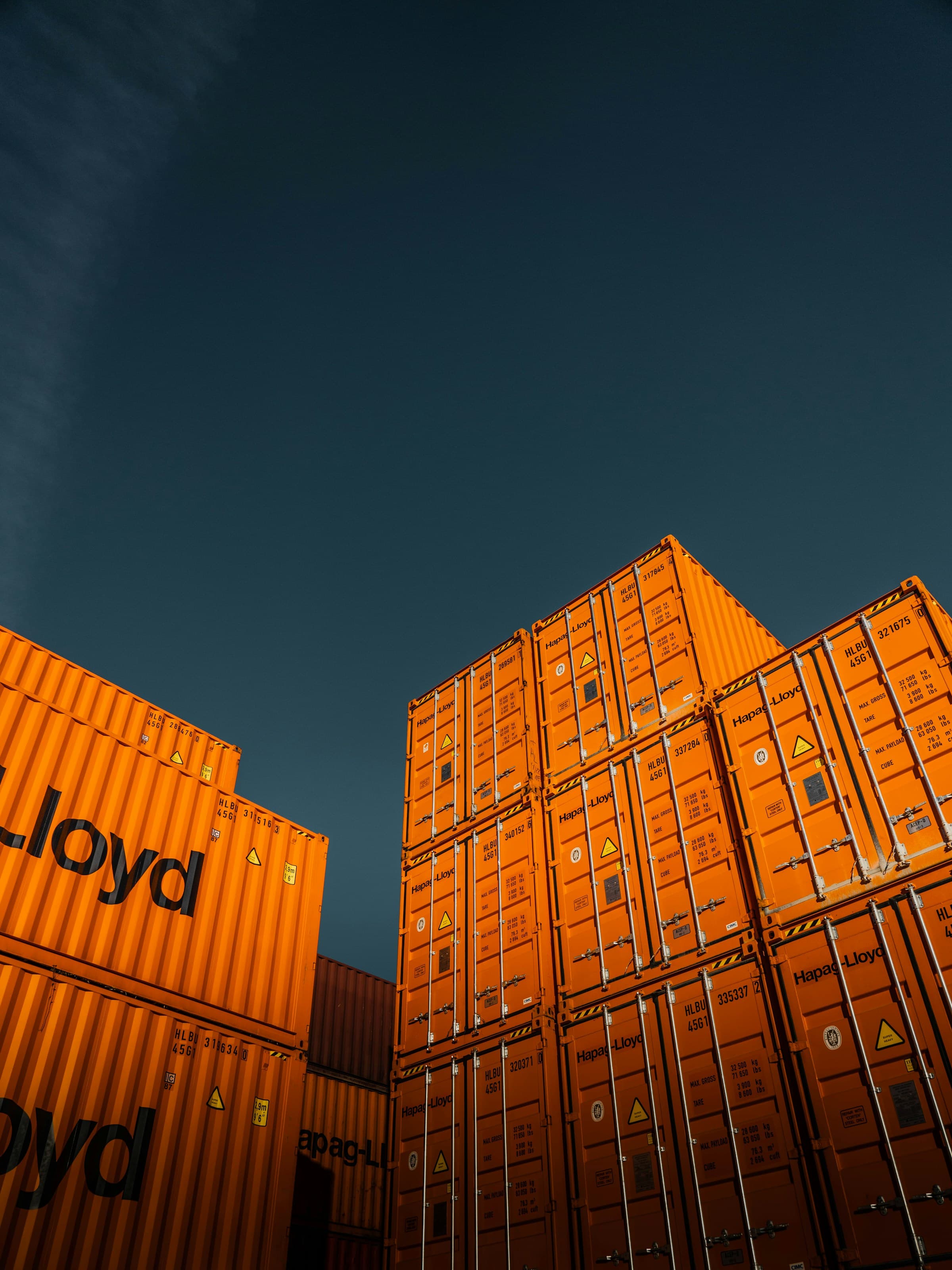 Stacked orange Hapag-Lloyd shipping containers rising against a dark blue evening sky